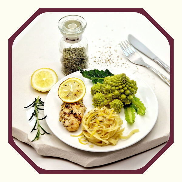 Rosmarin im Glas neben einem Teller mit einem Nudelnet, Romanesco, Rosmarin-Zitronen-Hähnchen und einer Scheibe Zitrone, einem Rosmarinzweig und Besteck