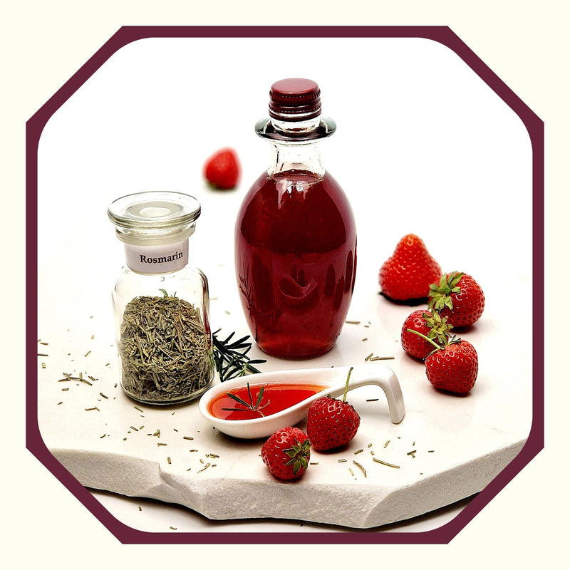 Rosmarin im Glas neben einer Flasche mit Erdbeer-Rosmarin-Essig, einem weißen Porzellanlöffel mit Essig, Erdbeeren, Rosmarin