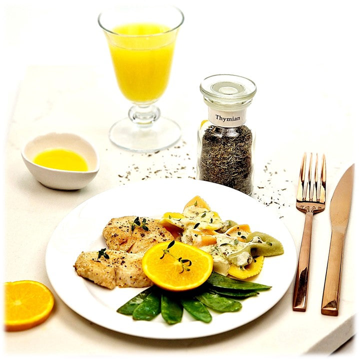 Thymian im Glas, ein Teller mit Hähnchenfilet, Nudeln, Zuckerschoten, Orange, daneben Besteck, eine Orangenscheibe, Orangensaft in einer kleinen Schale, ein Glas Orangensaft im Hintergrund