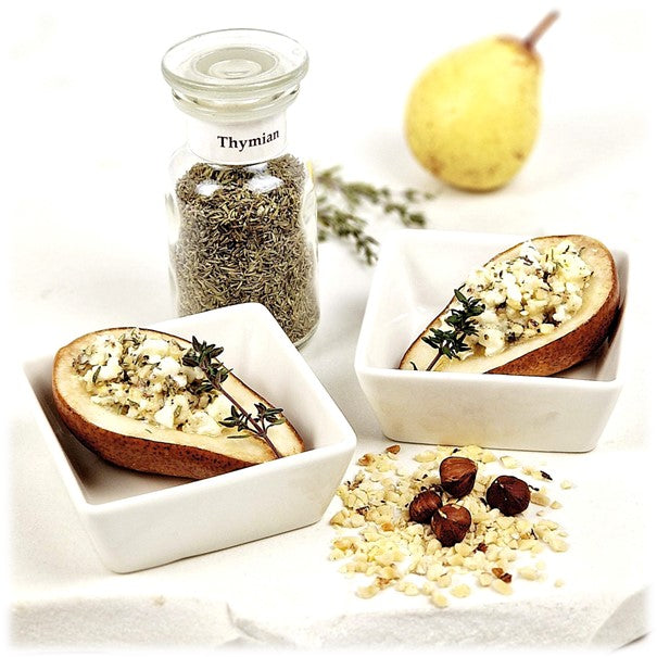 Thymian im Glas, daneben zwei viereckige Schalen mit jeweils einer Birnenhälfte, gefüllt mit einer Mischung aus Ziegenkäse, Thymian, Honig, Haselnüssen. Davor vier ganze Haselnüsse auf gehackten Haselnüssen, im Hintergrund eine Birne und Thymian-Zweige.