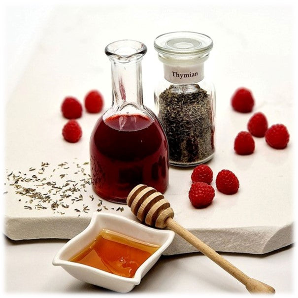 Thymian im Glas, neben einer kleinen Flasche mit Essig, im Hintergrund Himbeeren und Thymian, im Vordergrund eine viereckige Schale mit Honig und ein Honiglöffel aus Holz 