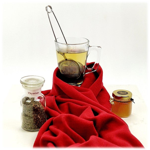 Thymian im Glas, daneben eine weiße Tasse Thymiantee, eingewickelt in einen Schal, daneben ein kleines Glas Honig