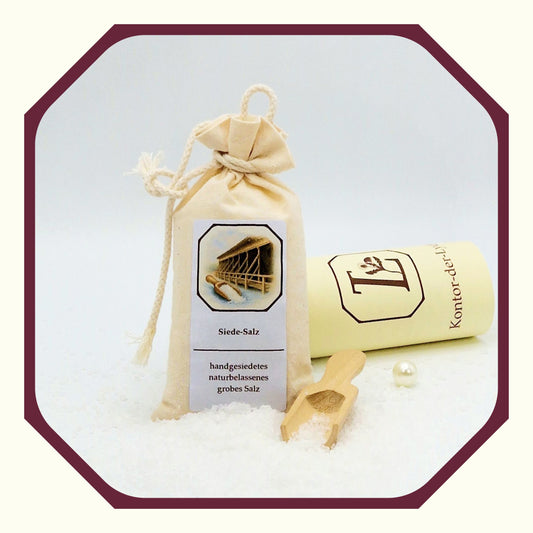 Eine Perle, eine rund gefaltete Karte mit schwarzem Schleifenband, ein Säckchen aus Leinen, gefüllt mit Siede-Salz, davor loses Siedesalz, ein kleine Salzschaufel aus Holz