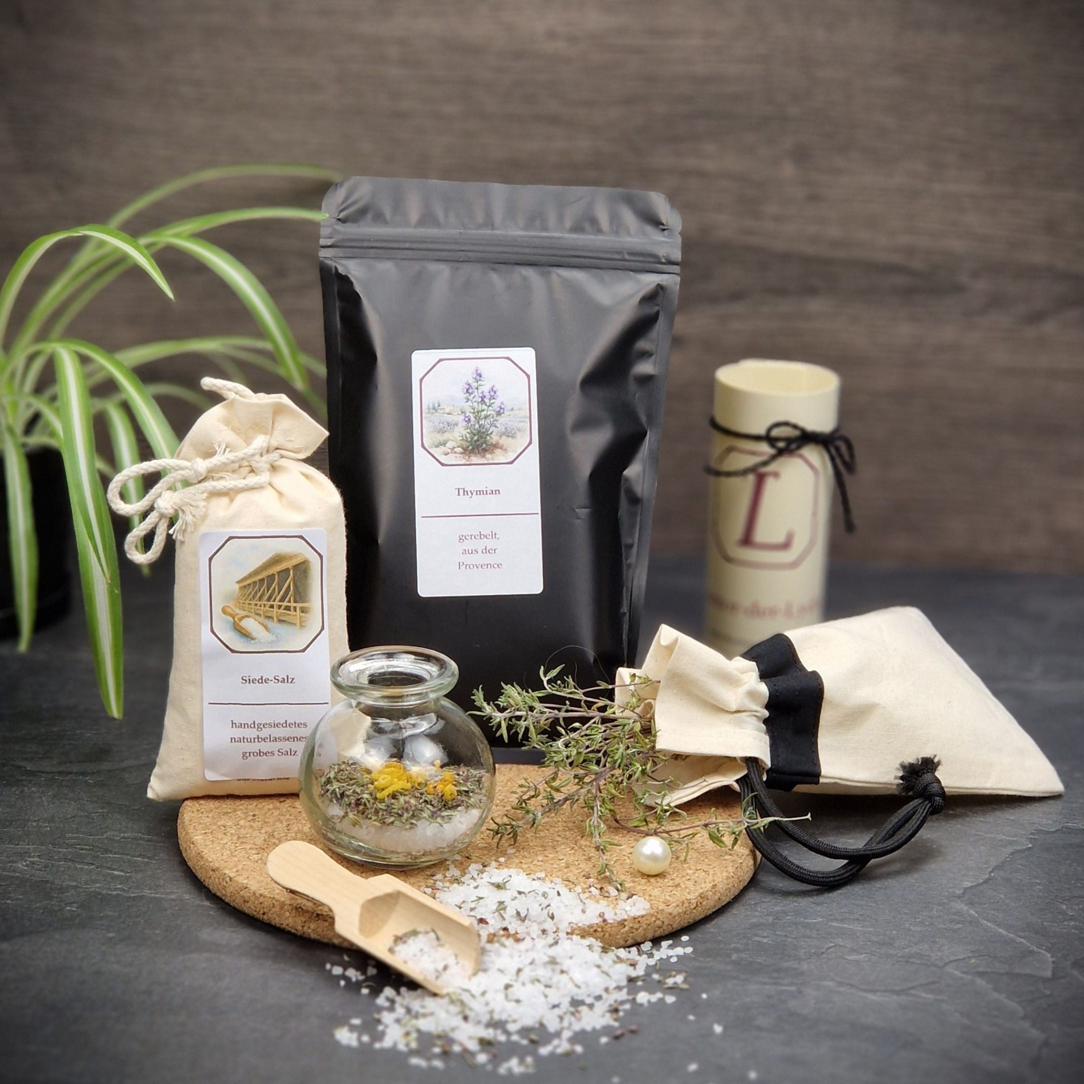 Auf einer Korkplatte: eine Klappkarte, eine Perle, eine Tüte mit Thymian, ein Salz-Sack, ein rundes Glas, gefüllt mit Salz, Thymian und Zitronenzesten, ein Kräutersäckchen mit Thymian, Thymian-Salz, eine Salz-Schaufel und eine Perle
