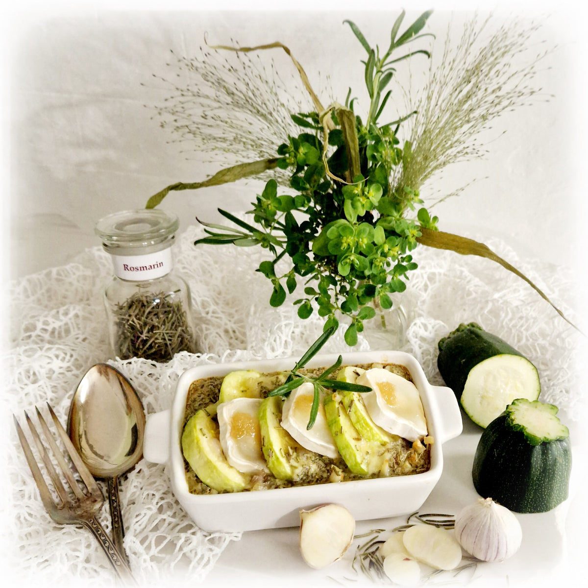 Stillleben mit einem Glas getrocknetem Rosmarin auf einer Spitzendecke mit Blumenstrauß, Rosmarin-Zucchini-Frittata mit Ziegenkäse und Honig, daneben Zucchini, Knoblauch, getrockneter Rosmarin und nostalgische Gabel und Löffel