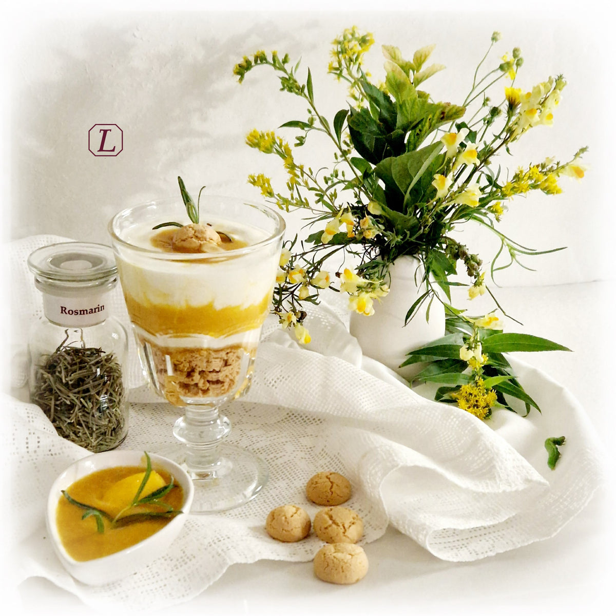 Stillleben mit einem Glas getrocknetem Rosmarin auf einer weißen Spitzendecke mit Blumenstrauß. Daneben ein elegantes Dessertglas mit geschichteten Amarettini, gelbem Pfirsichmus und cremigem Joghurt, garniert mit einem kleinen Gebäckstück und Rosmarin. Im Vordergrund befinden sich eine kleine weiße Schale mit Pfirsichmus und Rosmarin-Zweigen sowie vier Amarettini.